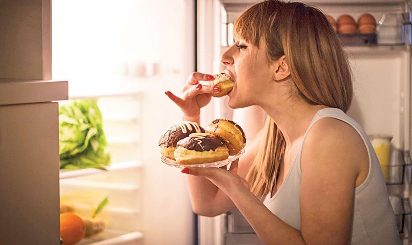 Close up image of a young woman with eating disorder, having a midnight snack - eating donuts, in front of the refrigerator.