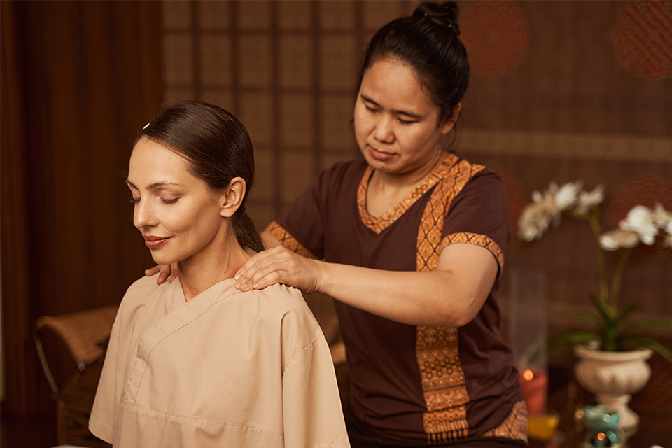 A woman therapist gently performs a shoulder massage on a relaxed client, with her eyes closed on a calm spa setting.