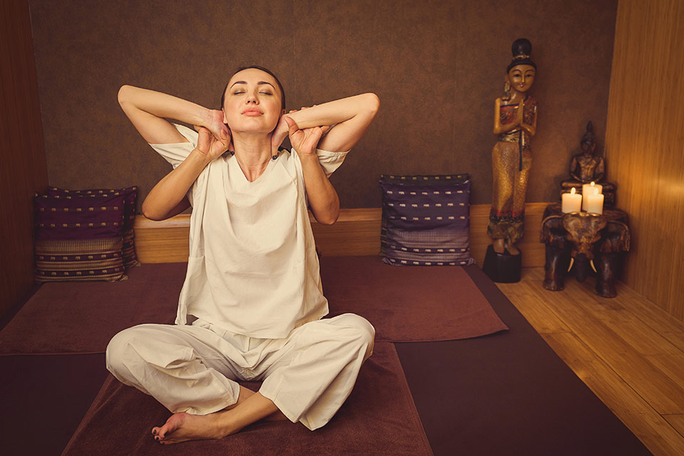 Woman sits cross-legged in a yoga pose, while therapist gently assists her with body stretching techniques on a mat on the floor.